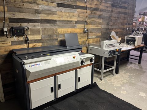Signstech glue binding machine, paper cutter and folder set up along a rustic wood wall